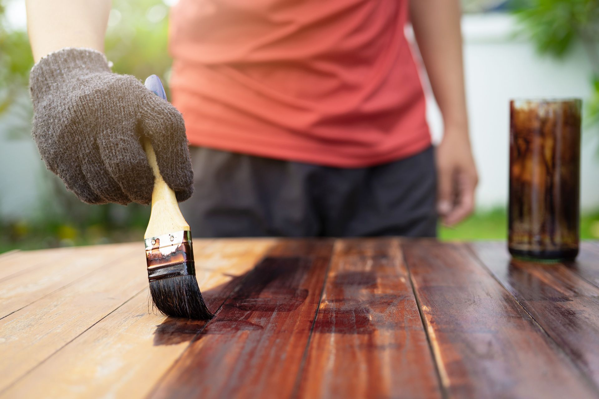 A person in a red shirt and work gloves uses a brush to apply dark wood stain to a wooden surface.