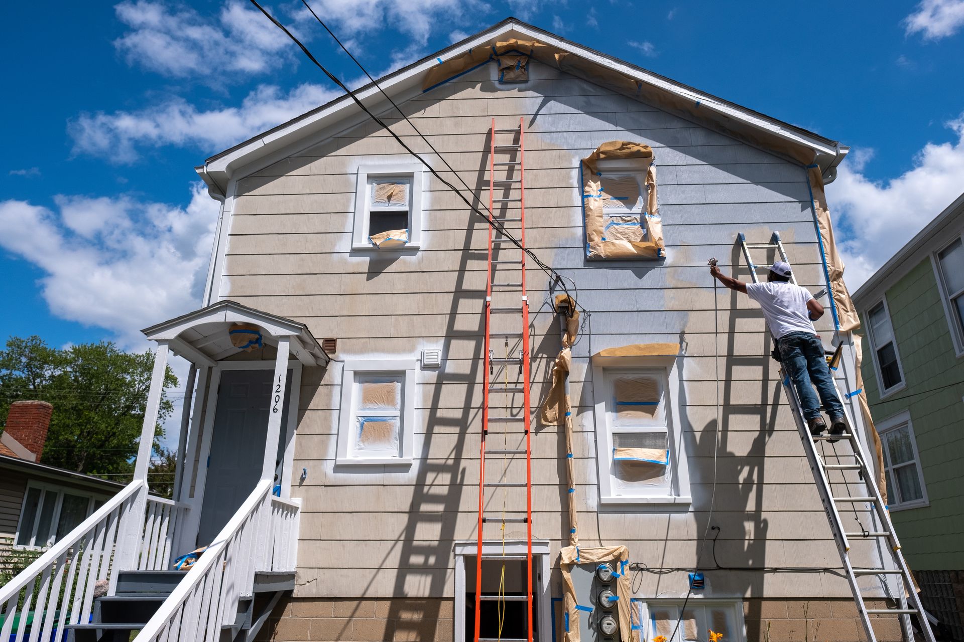 A worker on a ladder paints the side of a two-story house under a blue sky.