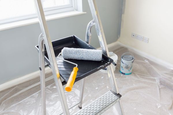 A metal step ladder holds a paint tray and roller, with a paint can nearby on a plastic-covered carpet in a room.