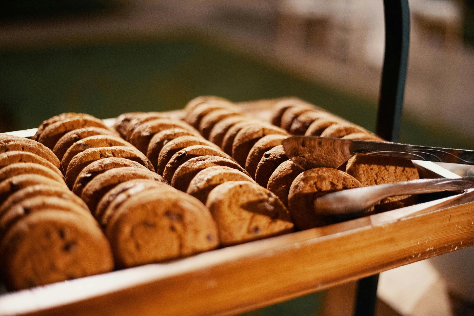 A wooden tray filled with chocolate chip cookies and tongs.
