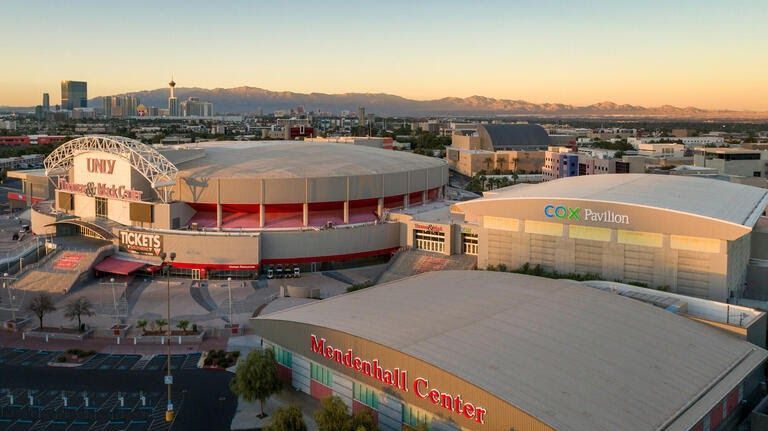 Aerial view of the UNLV campus with three buildings: Thomas & Mack Center, Cox Pavilion, and Mendenhall Center, with mountains in the background.