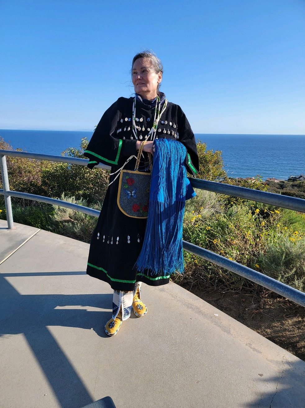 Person in black and green traditional clothing stands by a railing, holding a blue scarf with the ocean in the background.