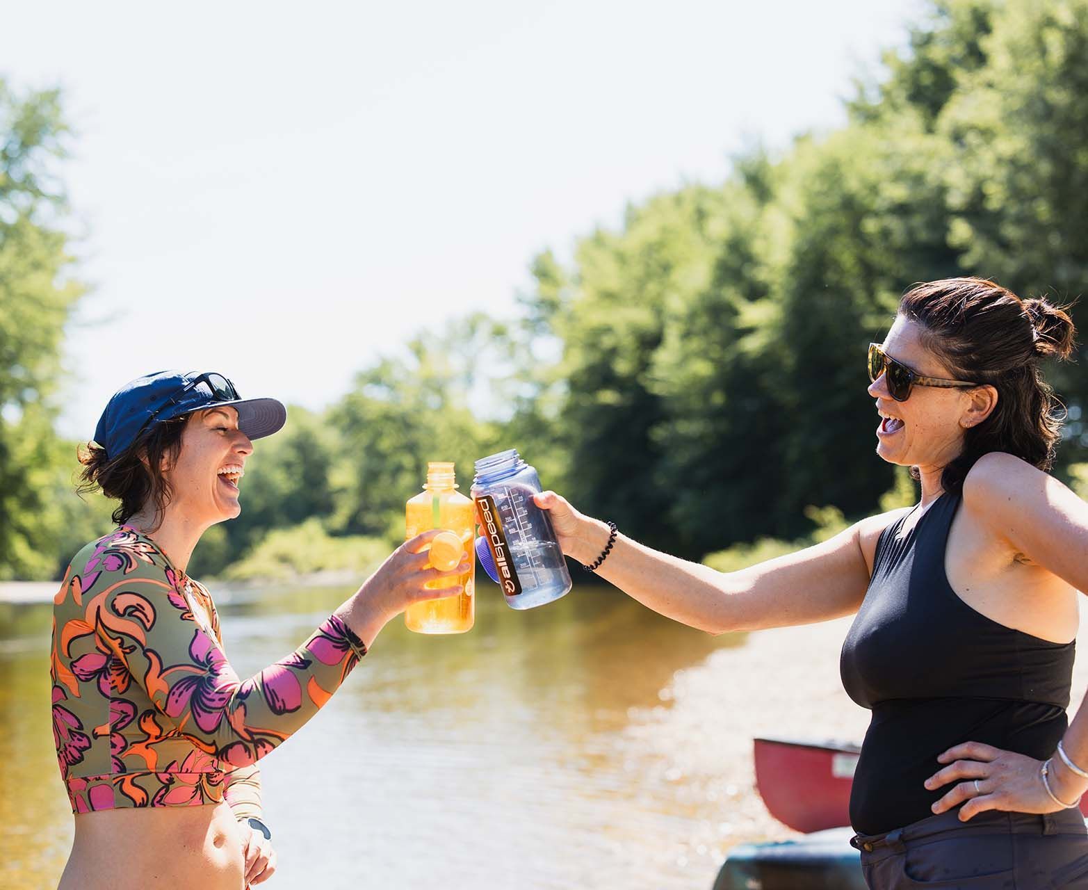 Two people on a boat toast drinks on a sunny river with trees in the background.