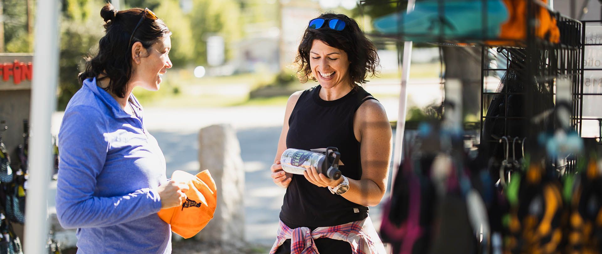 Two people talking outdoors by a market stall, one holding orange flowers and the other a water bottle.