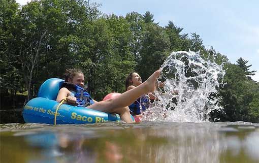 Two people in a blue inner tube splashing water on a lake with trees in the background