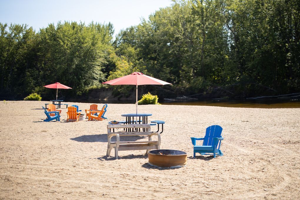 Sandy beach with Adirondack chairs, picnic table, and fire pit under red umbrellas near a river.
