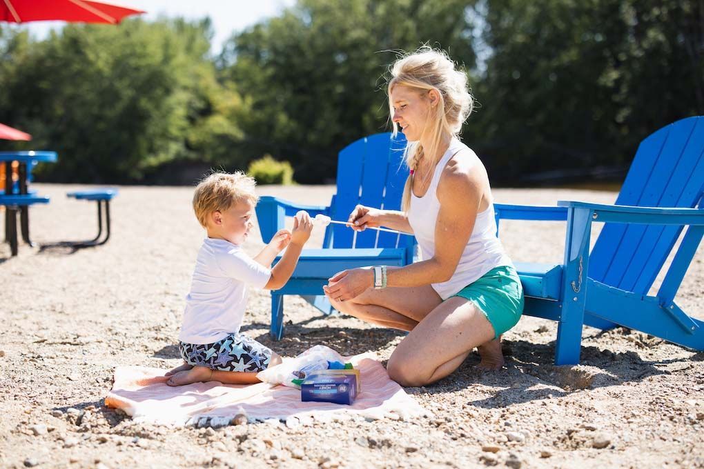 Woman applying sunscreen to a child's arm on a sandy beach; blue chairs and umbrella visible.