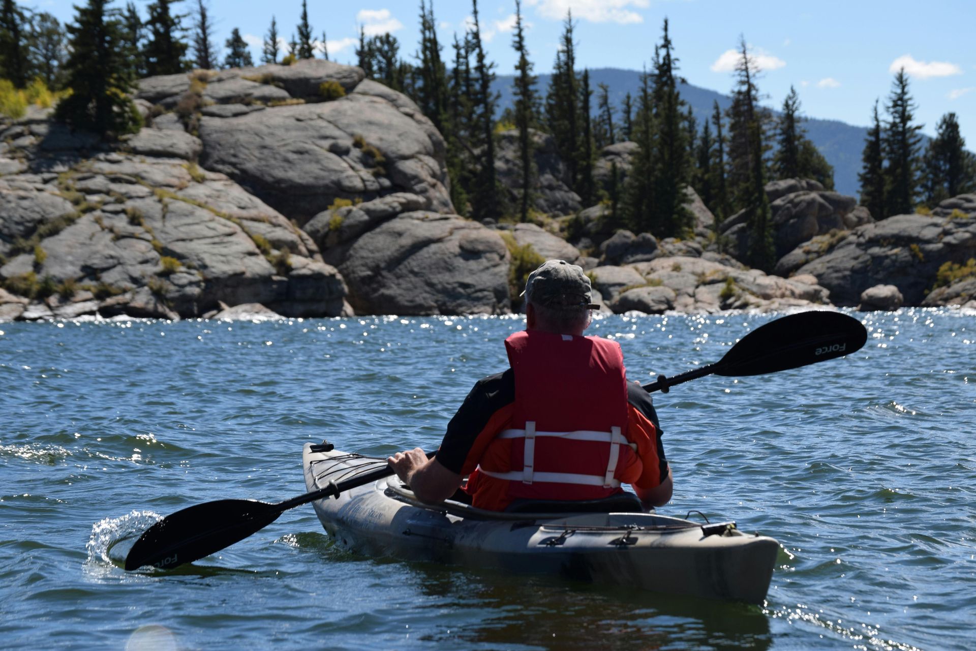 A person in a red life vest kayaks on a sunny, blue lake near rocky cliffs and pine trees.