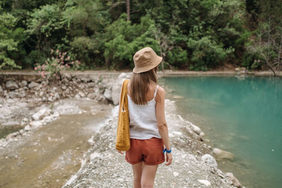 Person in a straw hat stands by a turquoise river beside a rocky shore and dense green trees.