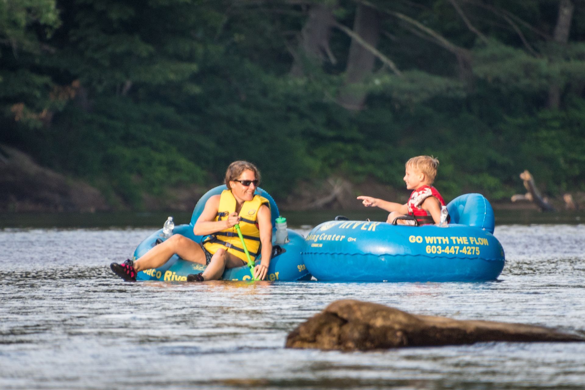 An adult and child in life jackets float in blue inner tubes on a river near a wooded shore.