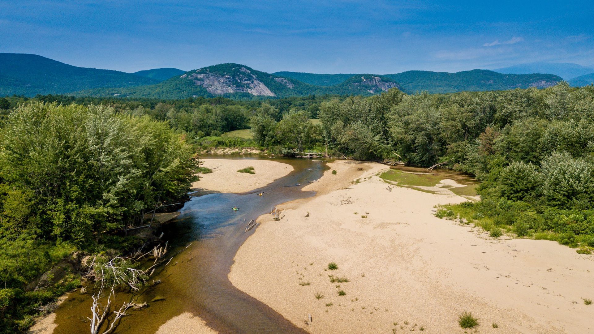 Aerial view of a river surrounded by trees and sand with mountains in the background