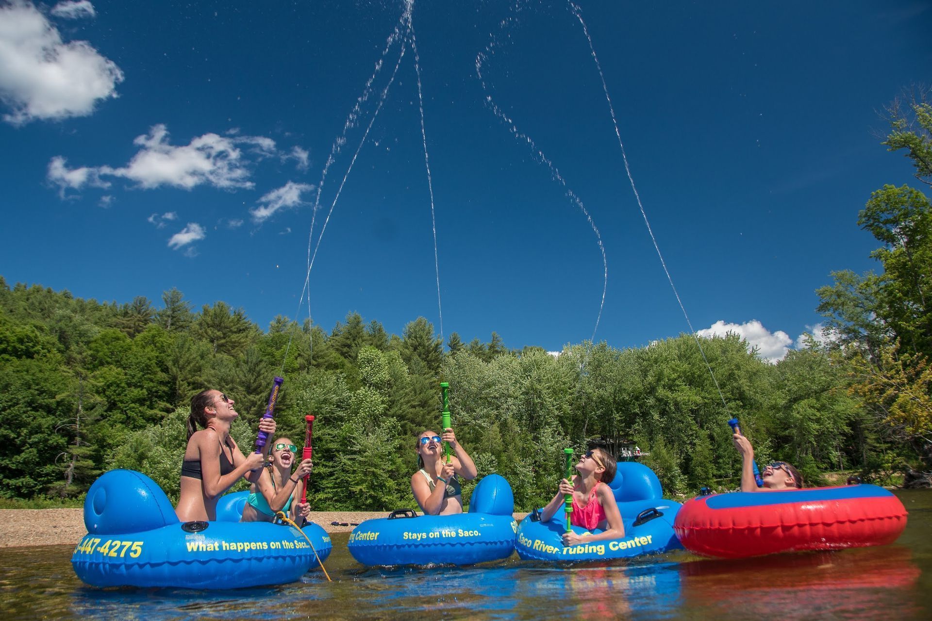 Four people in inner tubes float on a river, aiming water toys upward to create a spray against a bright blue sky.