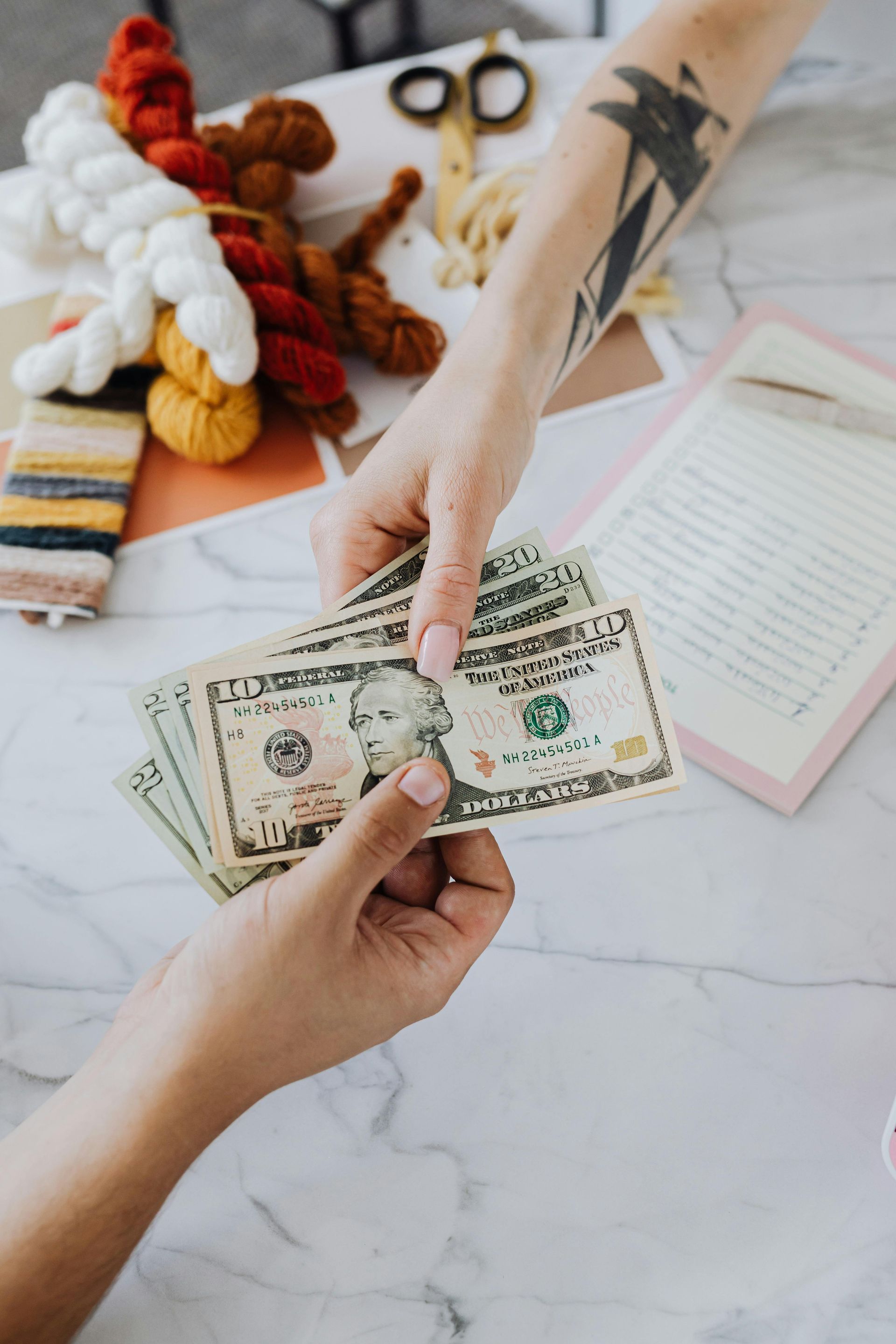 Hands exchanging cash over a marble surface with craft supplies in the background.