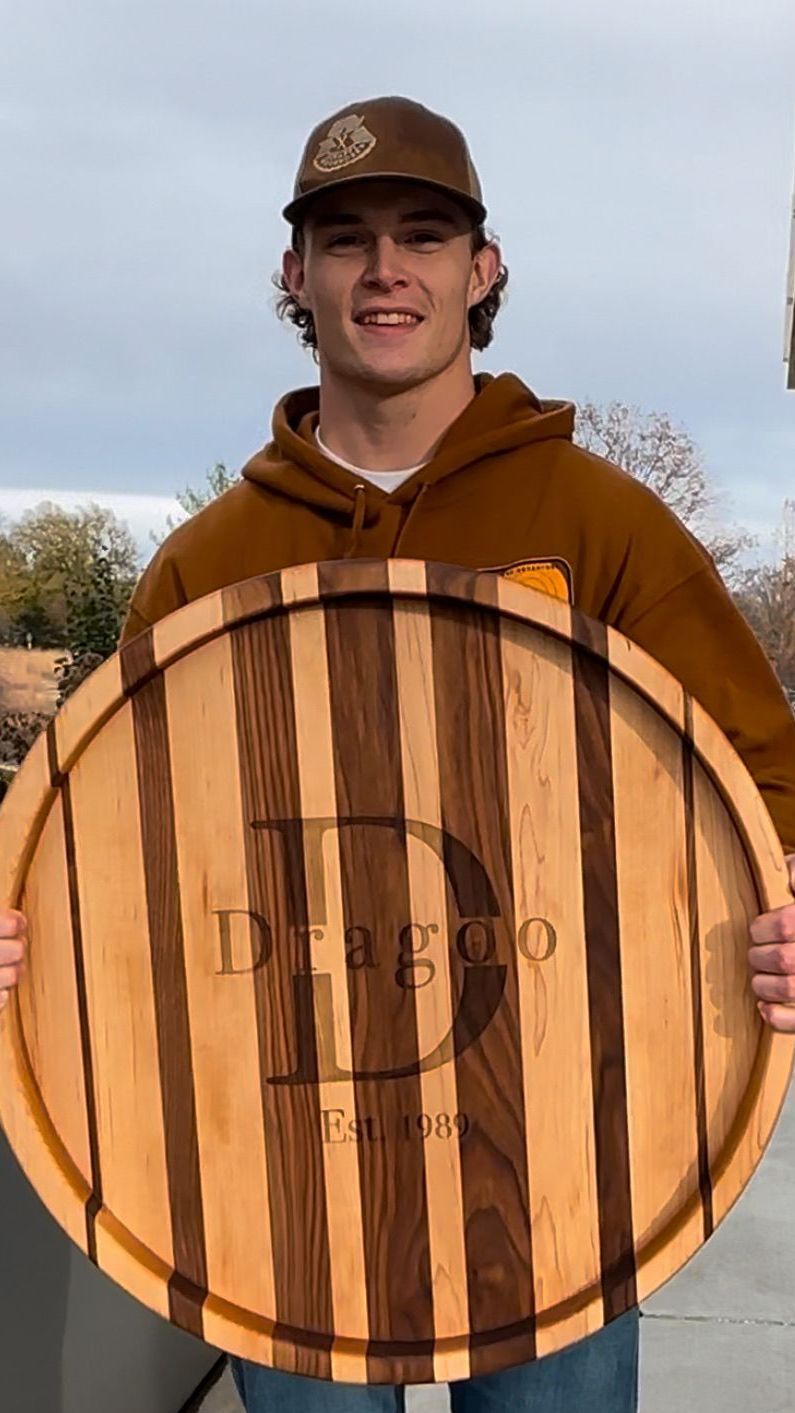 Man in brown holding a wooden platter with “Drago” engraved. Outdoors, smiling, wearing a cap.