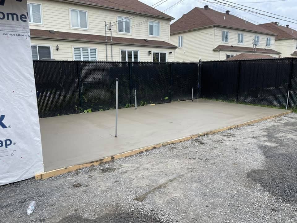 Terrasse en béton jouxtant une clôture noire et des maisons de banlieue, avec un poteau et du gravier au premier plan.