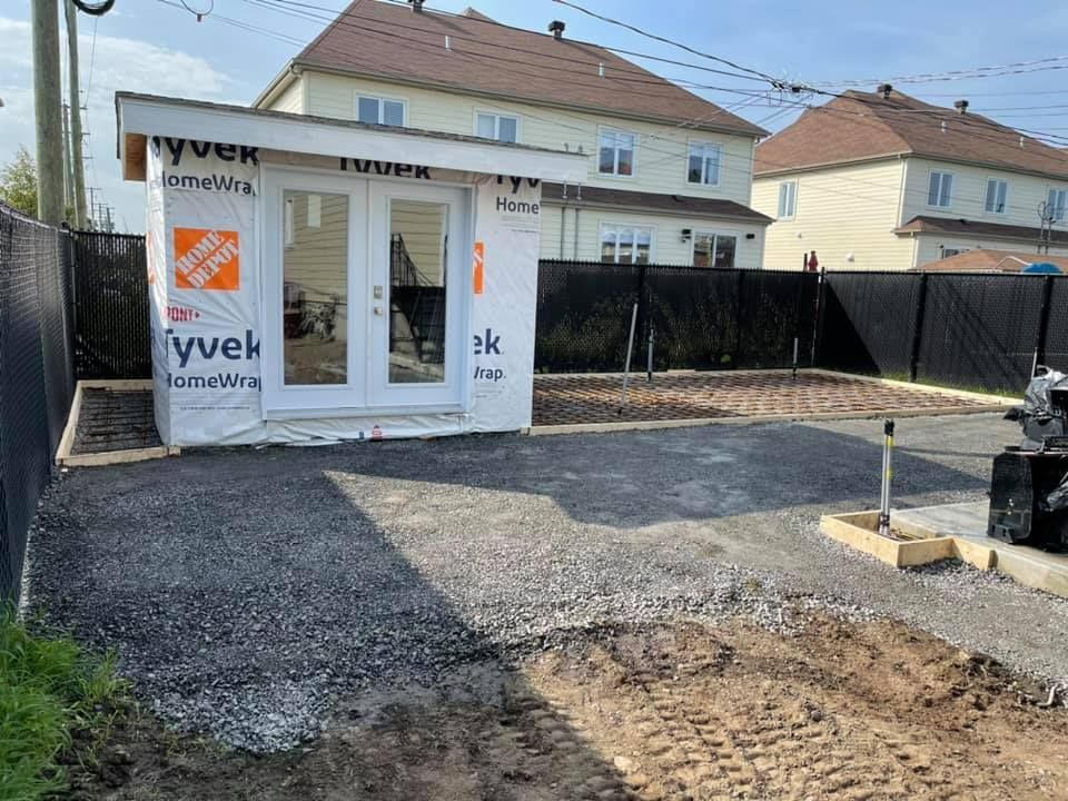 Maison en construction avec allée de gravier, façade blanche et clôture noire devant les maisons voisines.