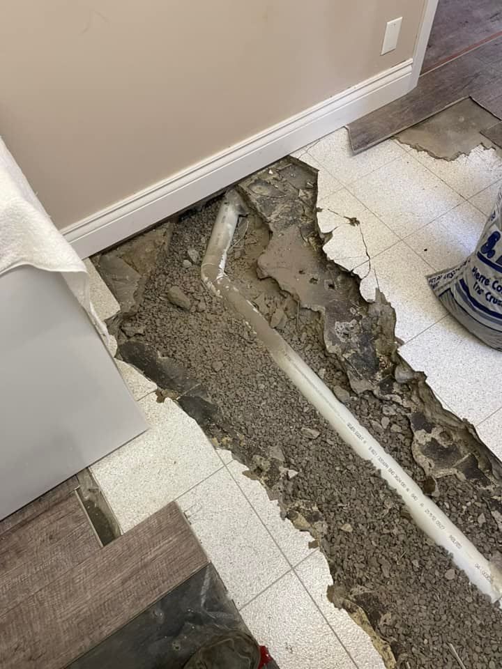 Sol de salle de bain endommagé, avec un grand trou laissant apparaître le béton et un tuyau blanc le long du carrelage.