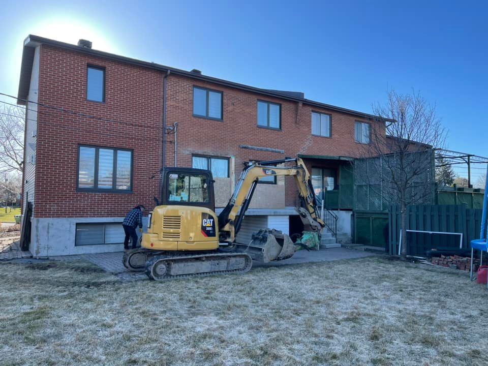 Une pelleteuse jaune est garée à côté d'une maison en briques en construction, avec une cour clôturée et un ciel bleu.