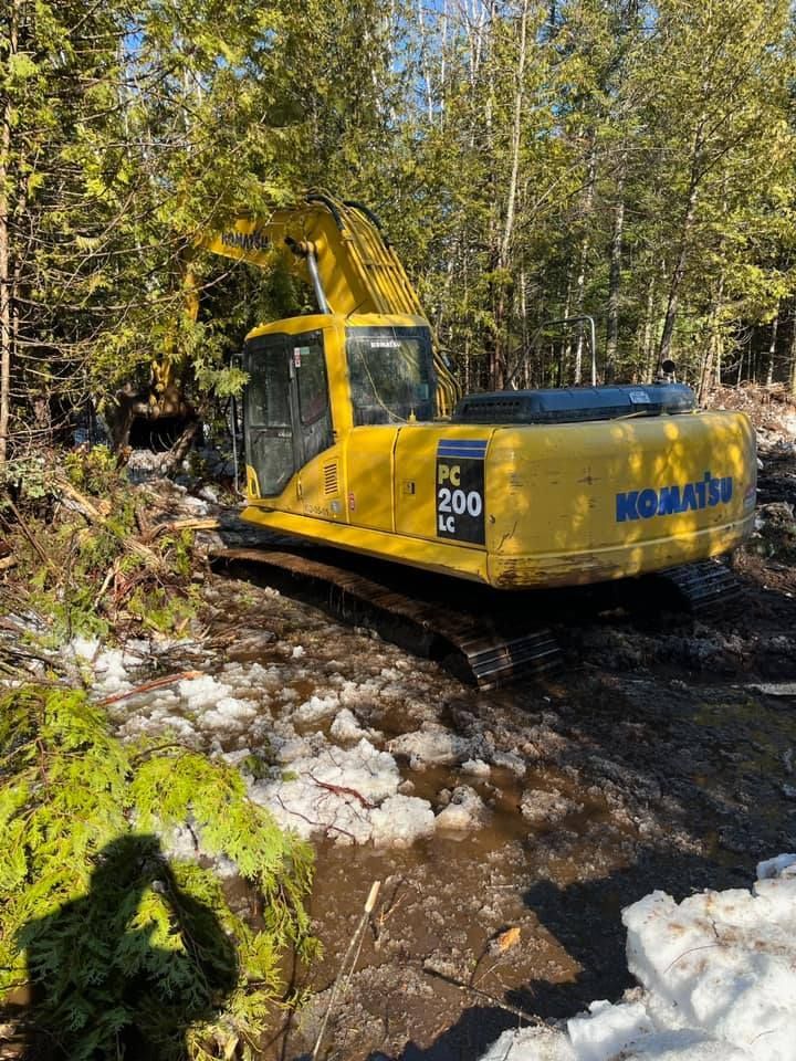 Une pelleteuse jaune traverse un ruisseau rocailleux dans une zone boisée.