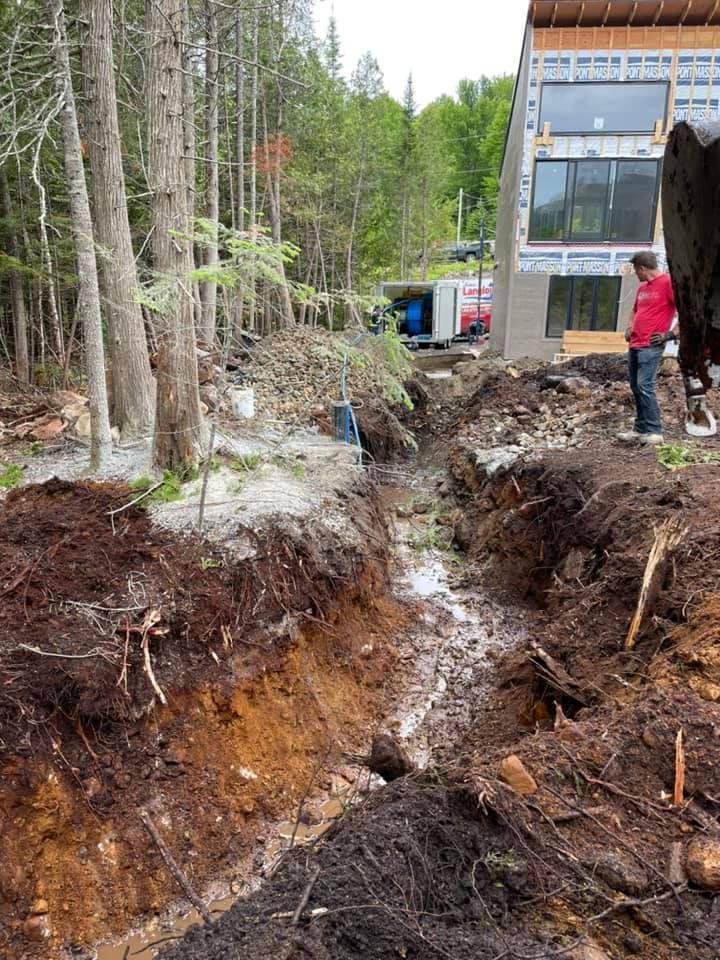 Tranchée profonde creusée à côté d'un bâtiment moderne dans une zone boisée, avec deux ouvriers à proximité.