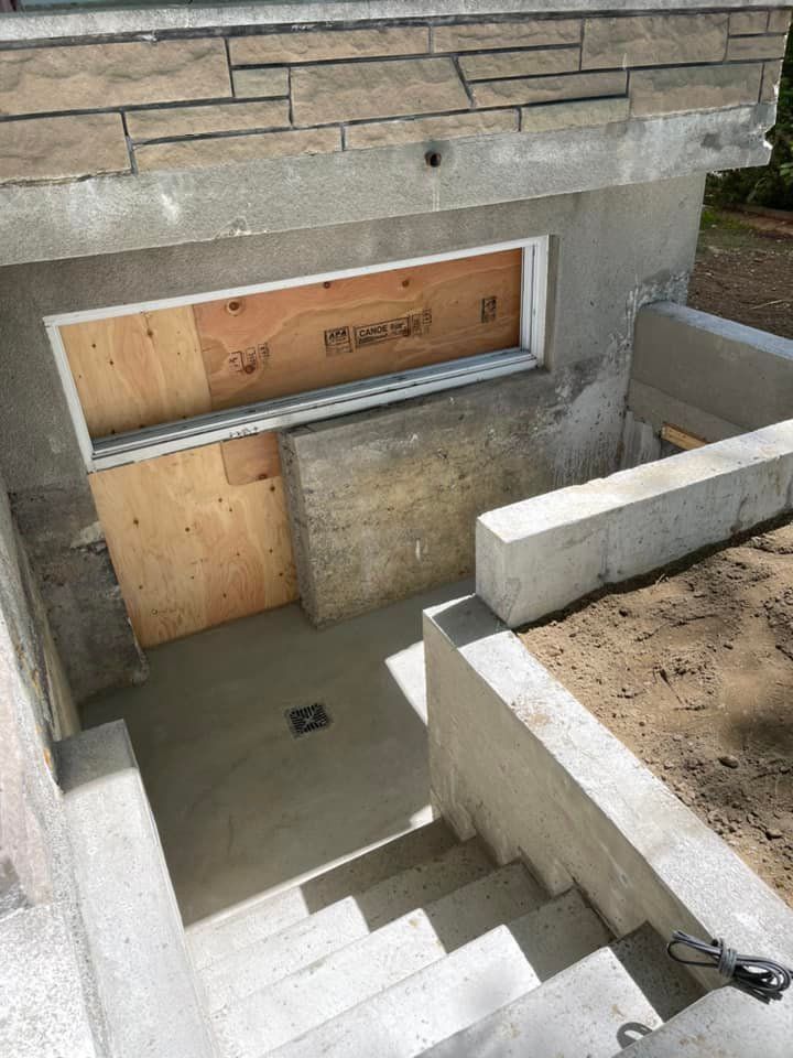 Entrée de sous-sol en béton avec escalier, rampe métallique et fenêtre condamnée sous un mur en pierre