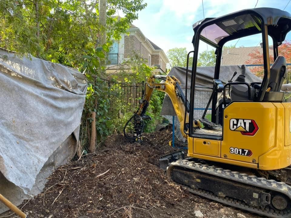 Une mini-pelle jaune CAT dégage les broussailles le long d'une clôture recouverte d'une bâche dans une ruelle étroite à l'arrière d'une maison.