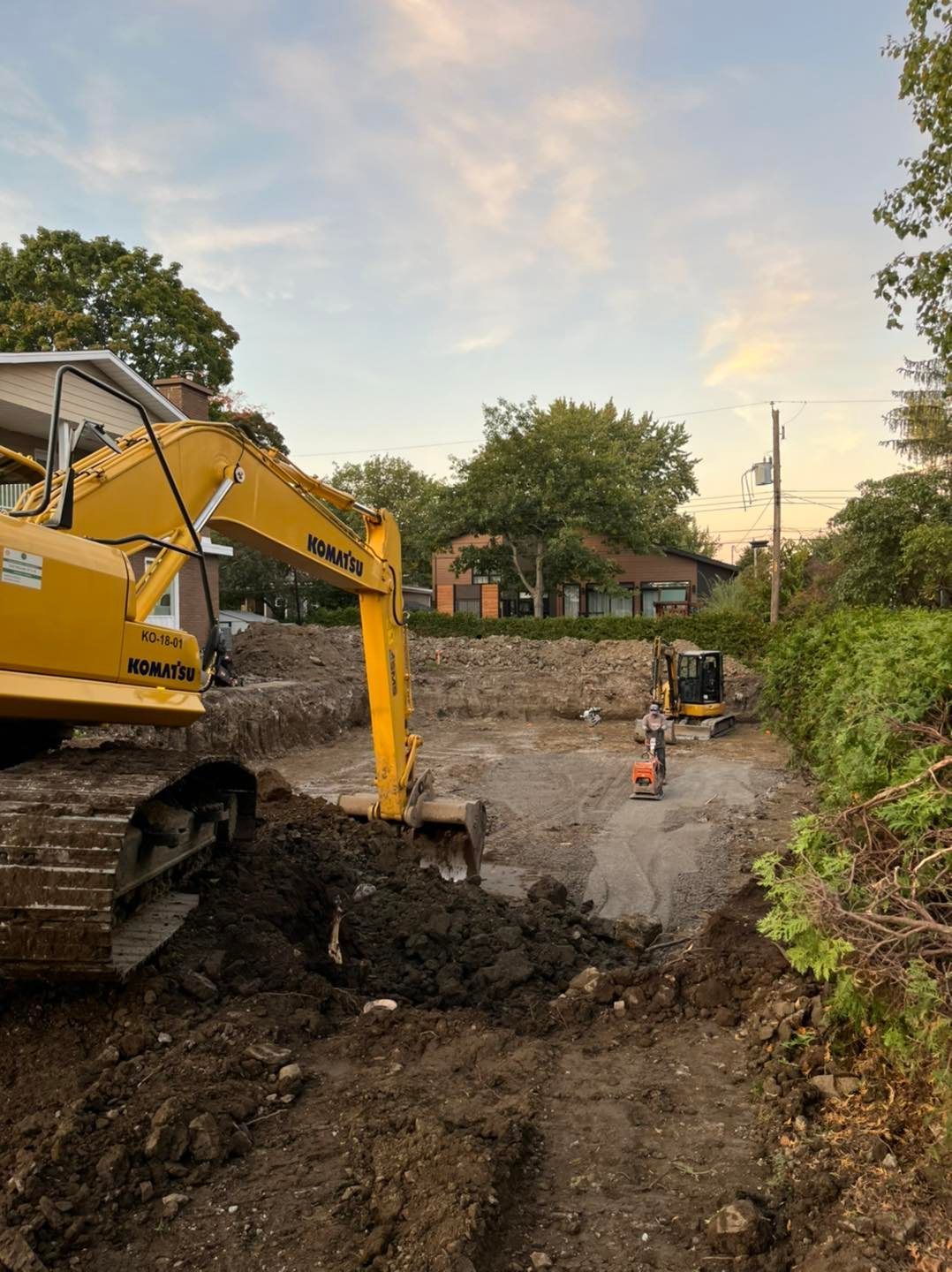 Une pelleteuse jaune creuse une tranchée sur un chantier boueux au crépuscule.