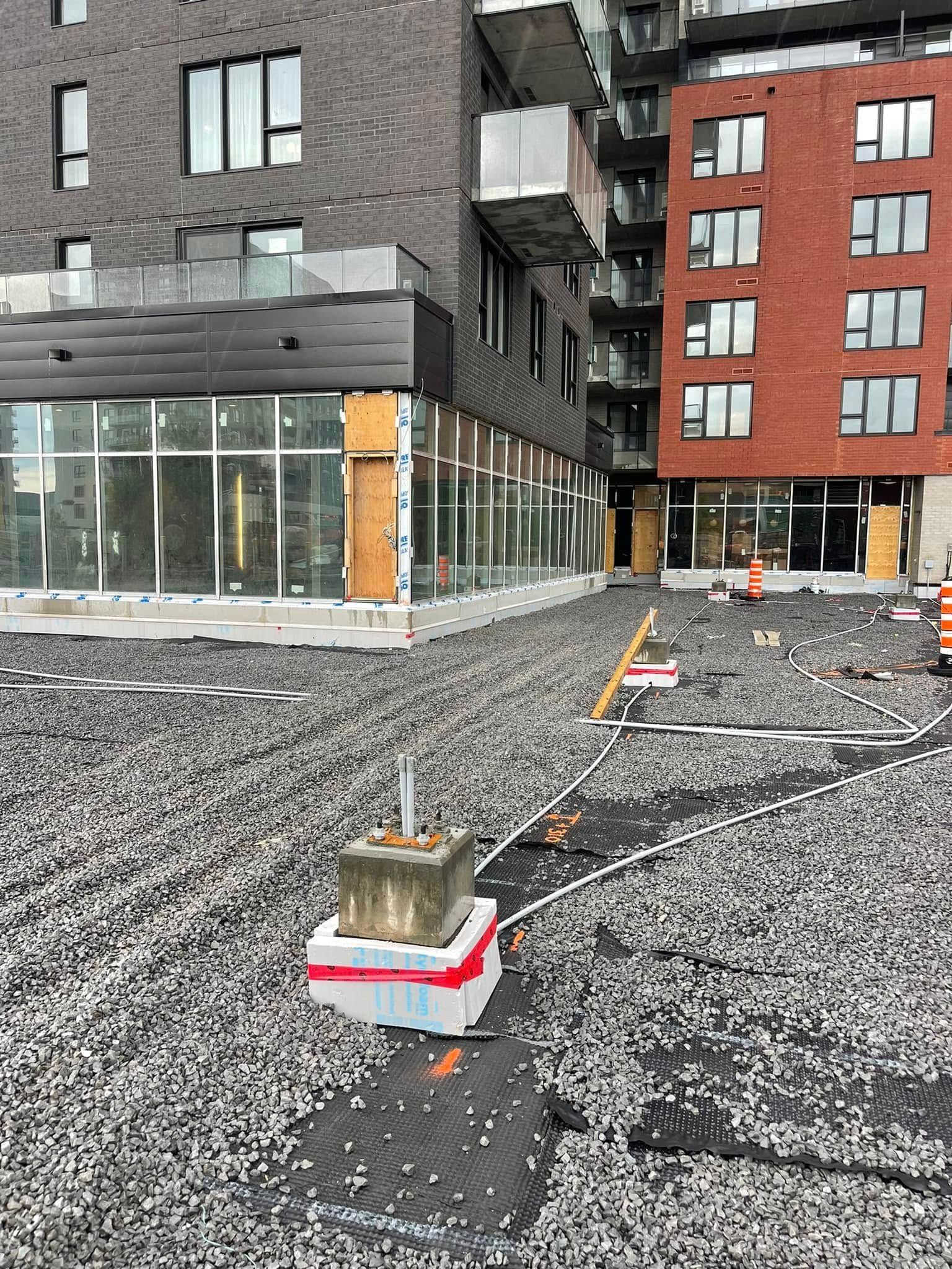 Chantier de construction extérieur à côté d'immeubles d'appartements, avec du gravier, un chemin balisé et des barrières temporaires.