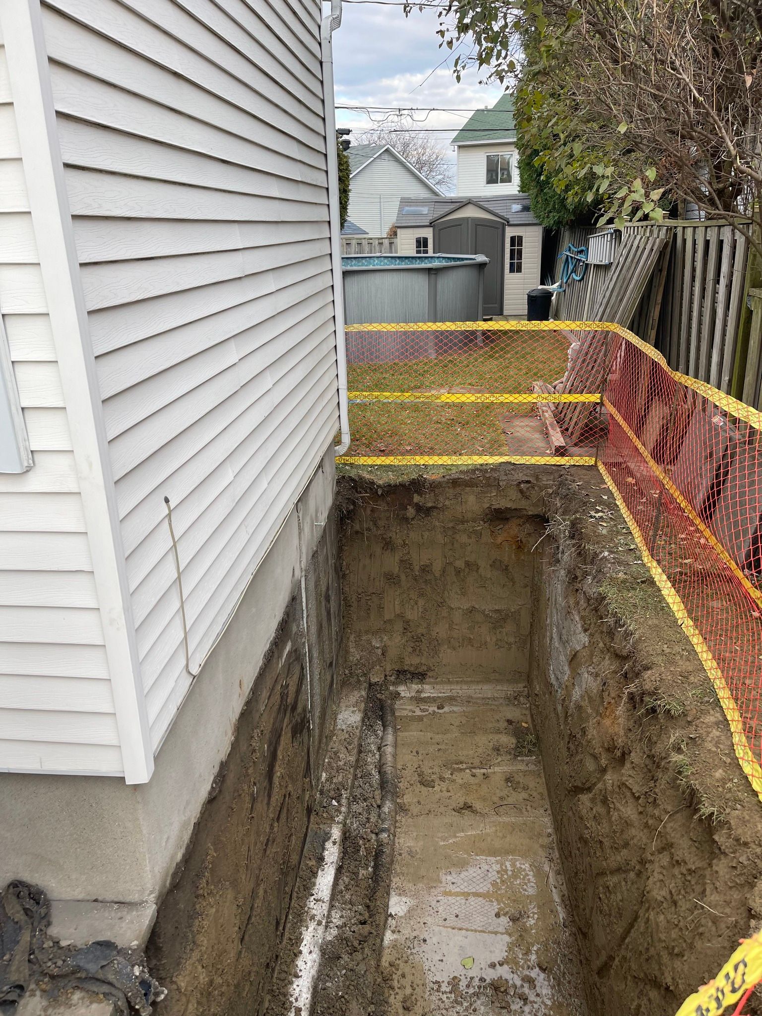 Étroite excavation jouxtant une maison, avec sol à nu et clôture de sécurité orange le long d'une cour arrière clôturée.
