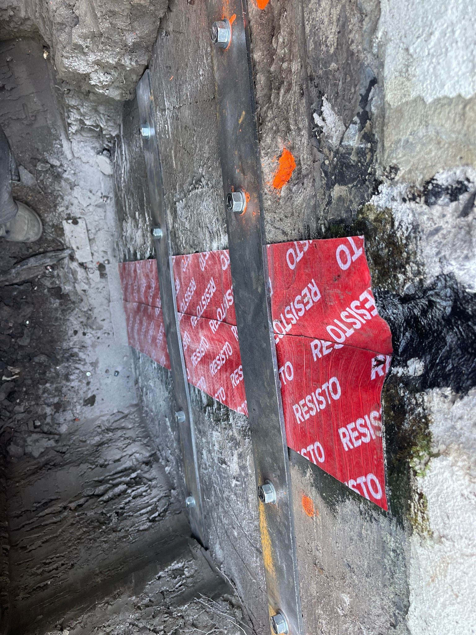 Patchs de réparation de chantier rouges et blancs sur un mur en béton gris rugueux, avec des boulons et des marques de repérage orange.