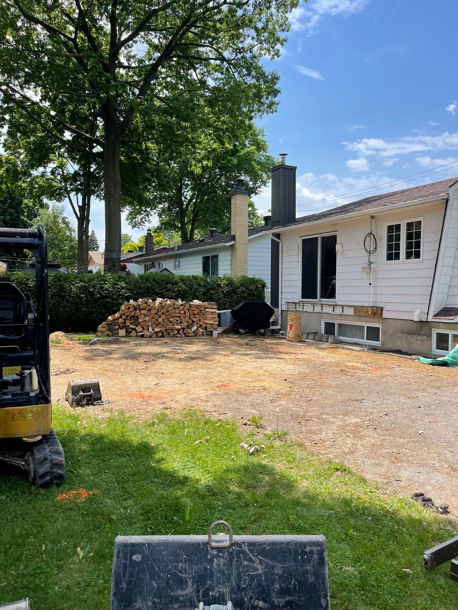 Rénovation d'un jardin avec une maison blanche, une zone de gravier, du bois empilé et une petite excavatrice à proximité.