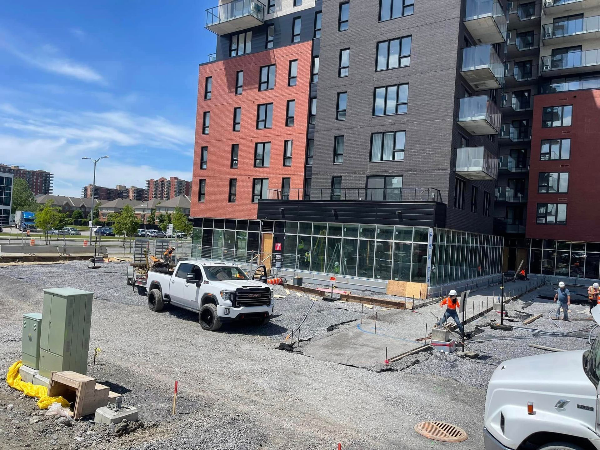 Chantier de construction jouxtant un immeuble d'appartements rouge et gris, avec des camions, du gravier et des ouvriers à proximité.