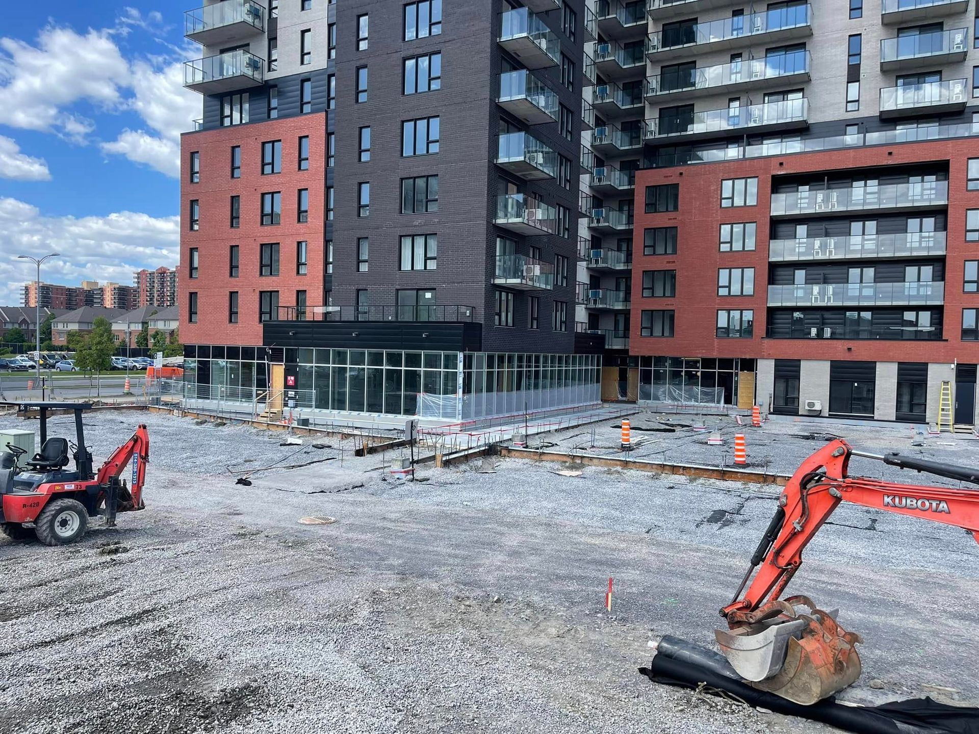 Chantier de construction devant un immeuble d'appartements en briques rouges, avec des excavatrices et un sol gravillonné.