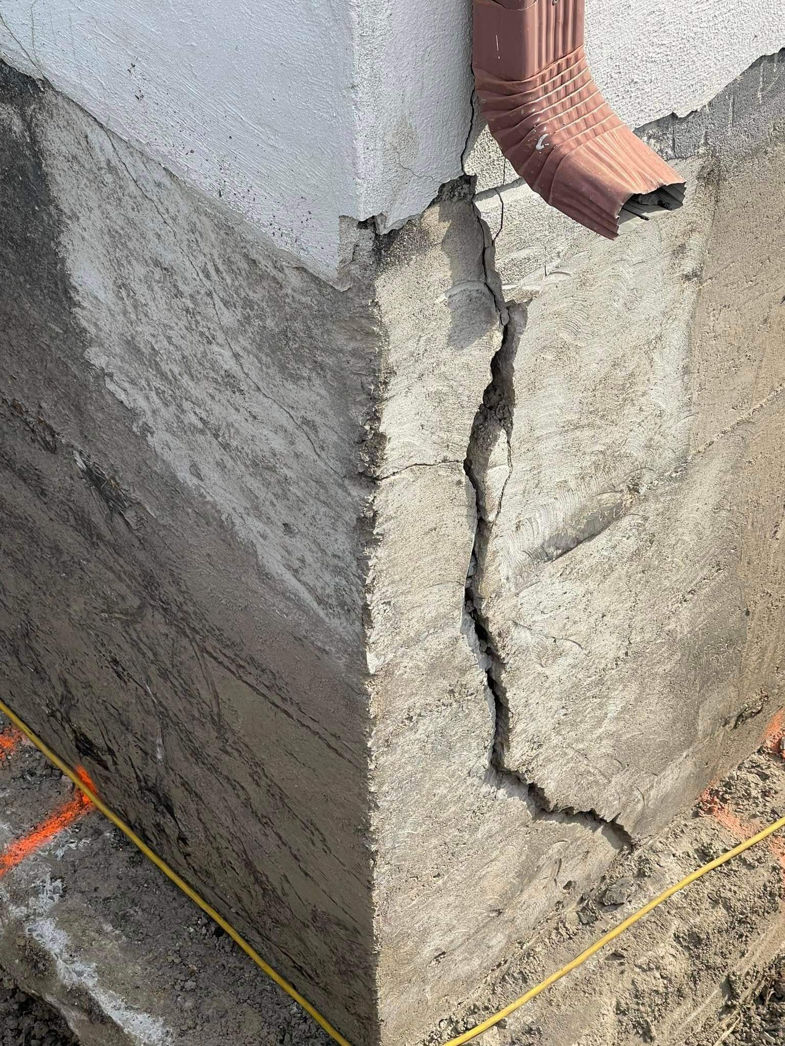 Coin de mur en béton fissuré, présentant des dommages apparents et une descente de gouttière rouge en haut.