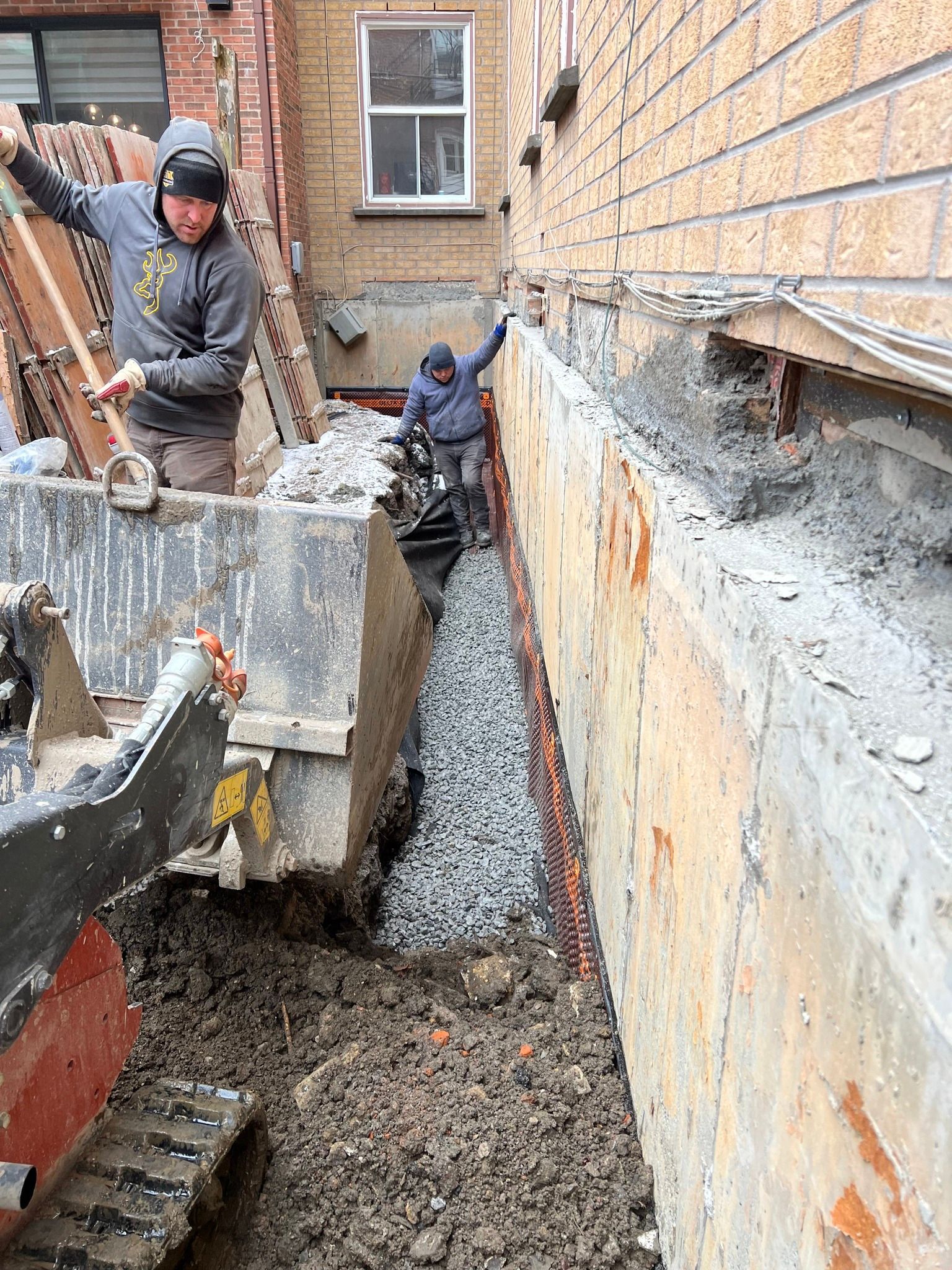 Des ouvriers coulent du béton dans une tranchée étroite à côté d'un bâtiment en briques, une personne supervisant le coulage.