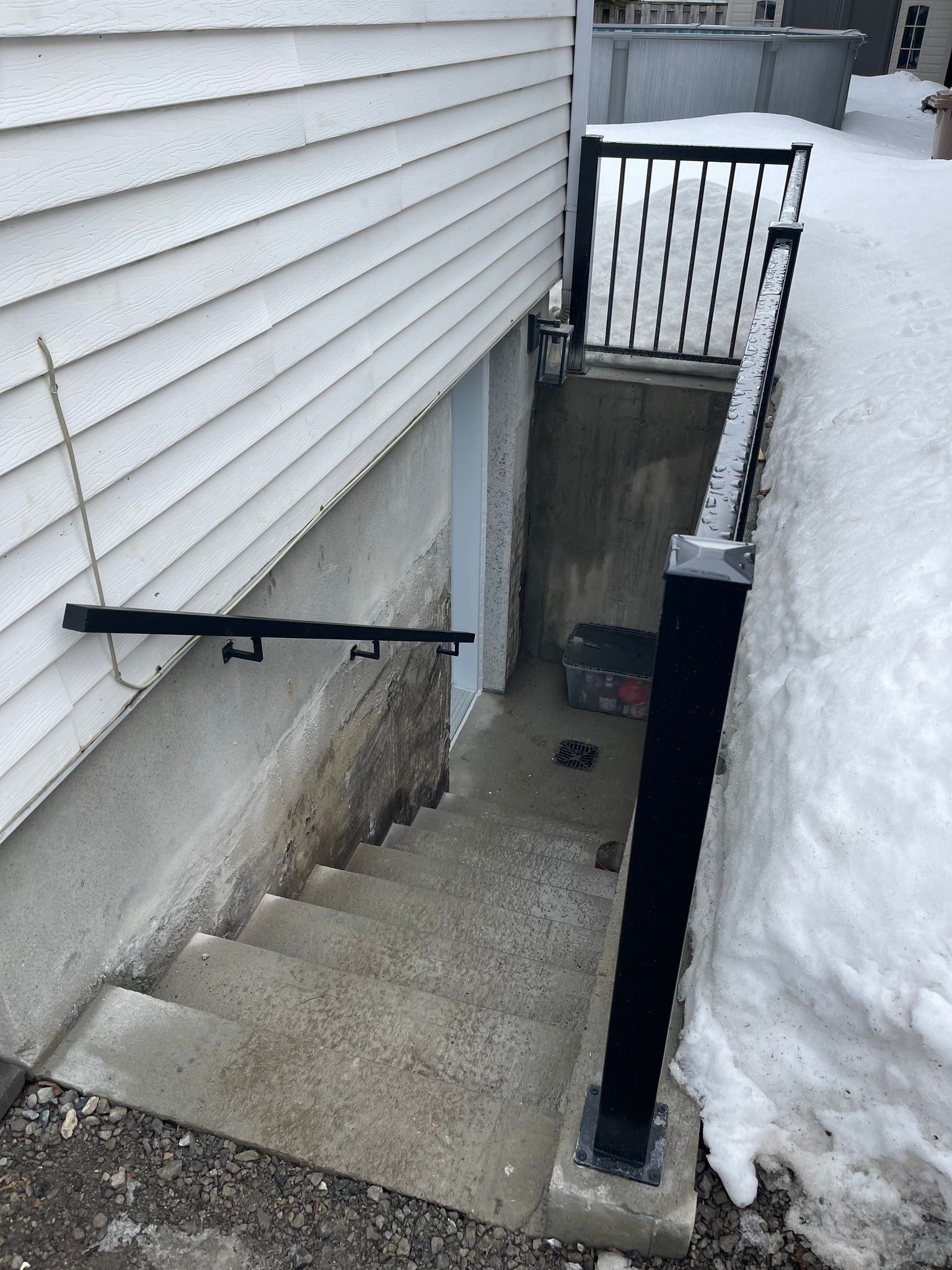 Escalier extérieur enneigé menant au sous-sol, à côté de la maison, avec une rampe noire et une grille métallique en bas.