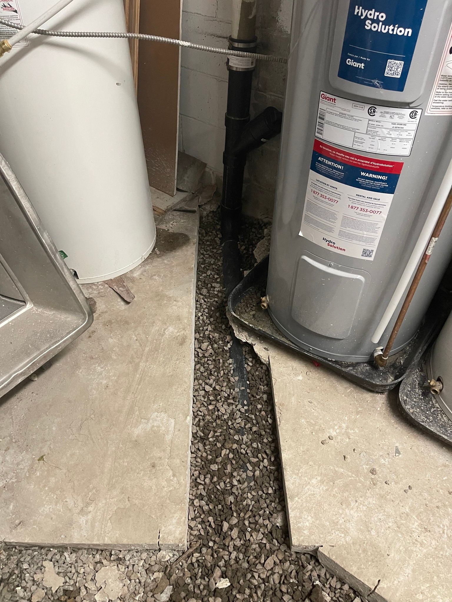 Tranchée ouverte creusée dans le sol en béton à côté d'un chauffe-eau et d'un tuyau blanc dans une buanderie