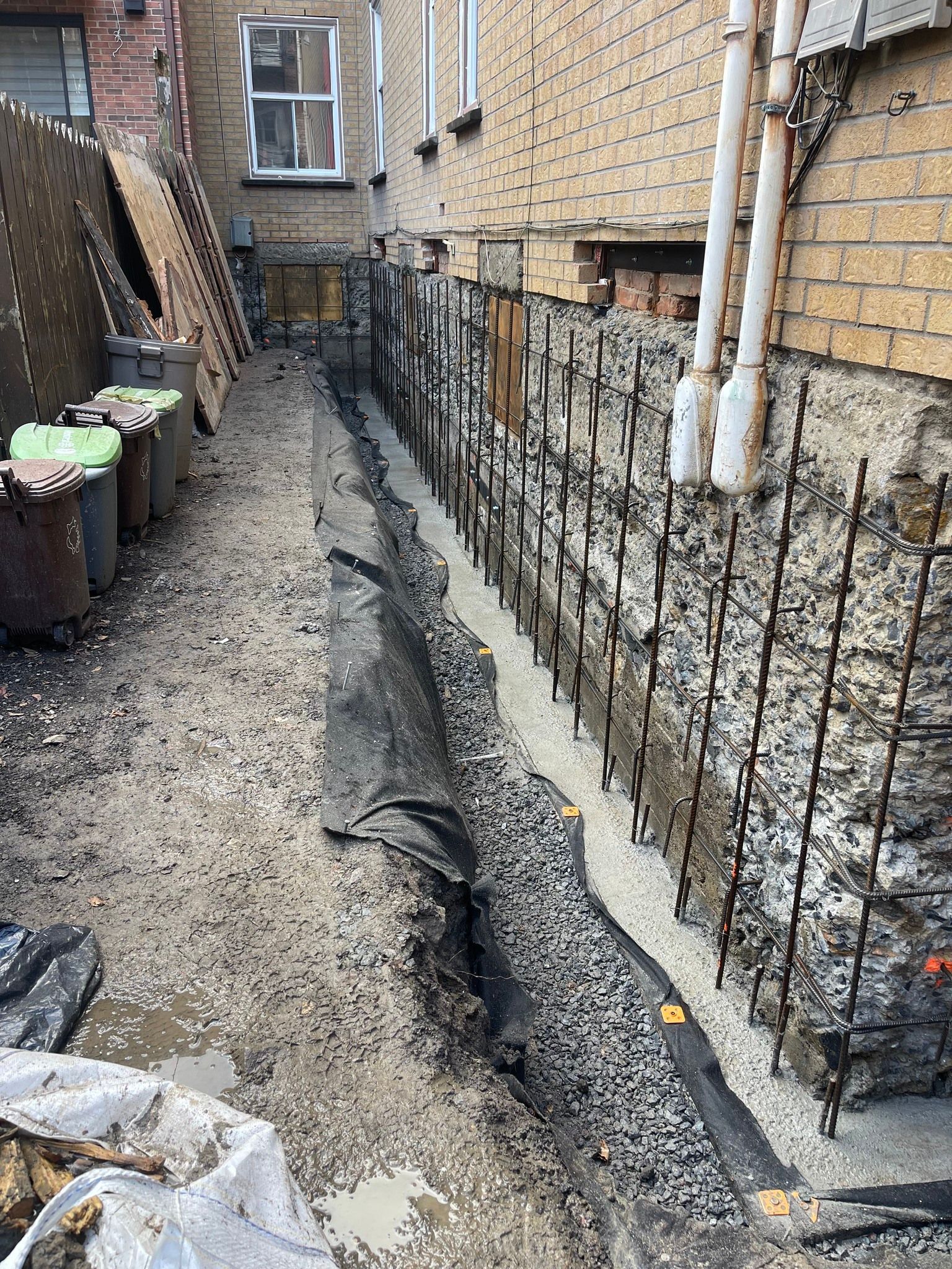 Allée étroite en gravier longeant un bâtiment en briques avec une tranchée et des canalisations.