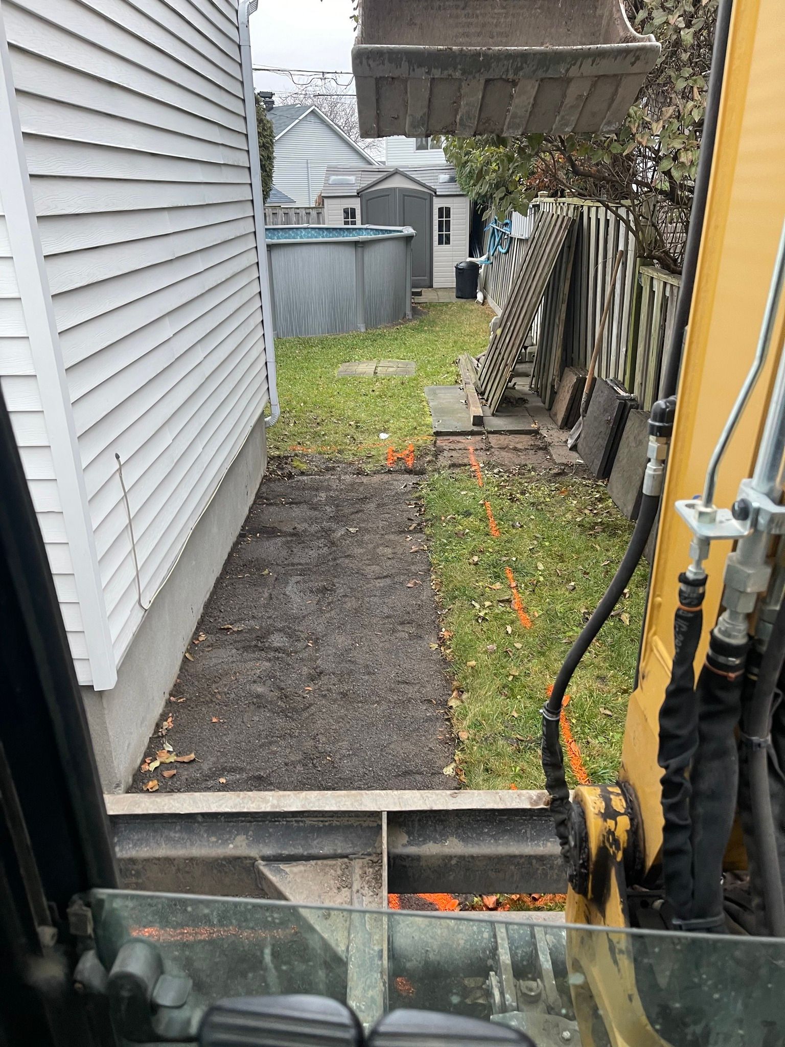 Passage étroit dans une cour arrière avec chemin de gravier, bardage blanc, clôture et bras d'excavatrice jaune au premier plan