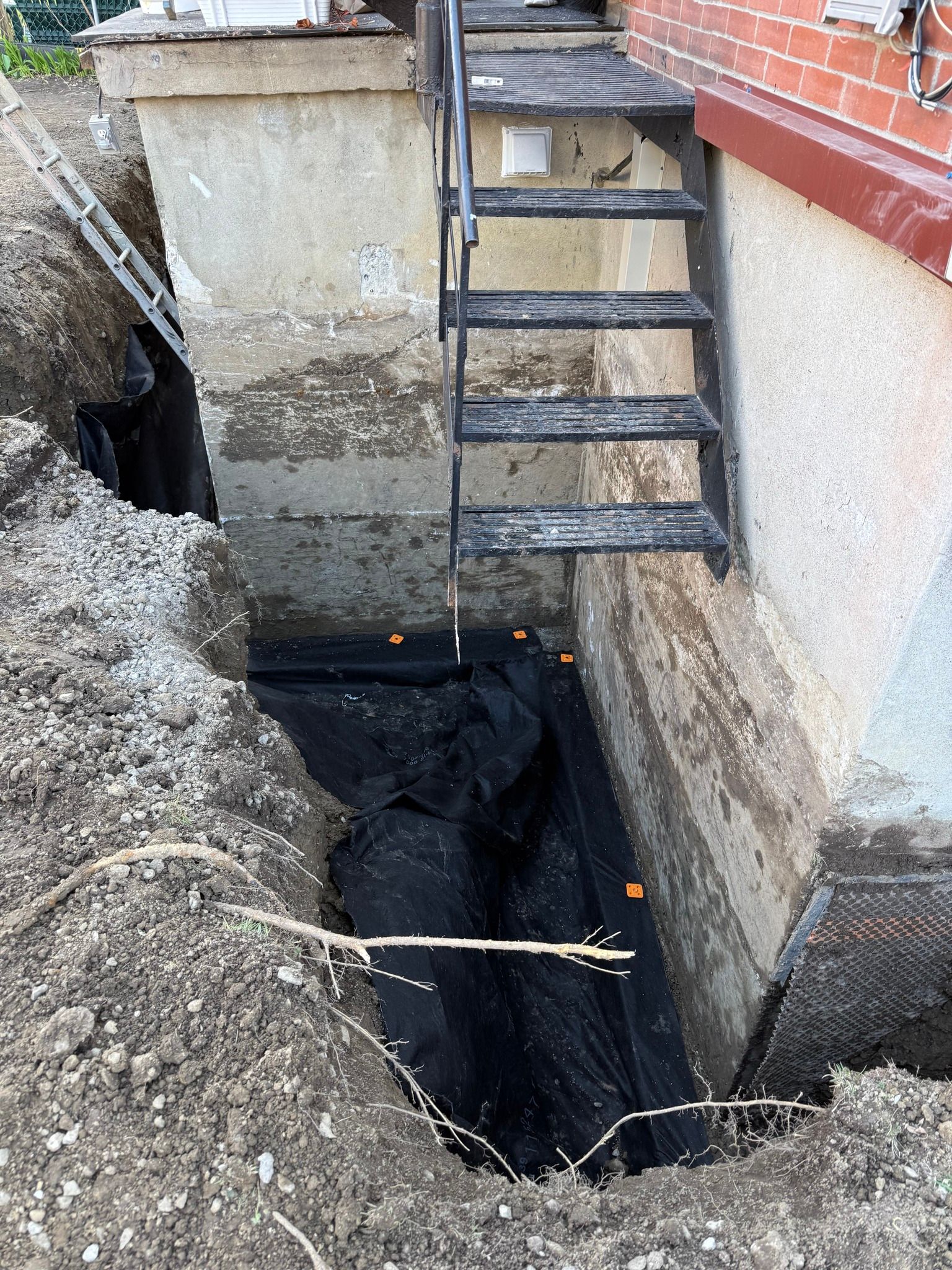 Fosse d'excavation à côté d'un mur de briques, avec une échelle métallique appuyée contre la tranchée.