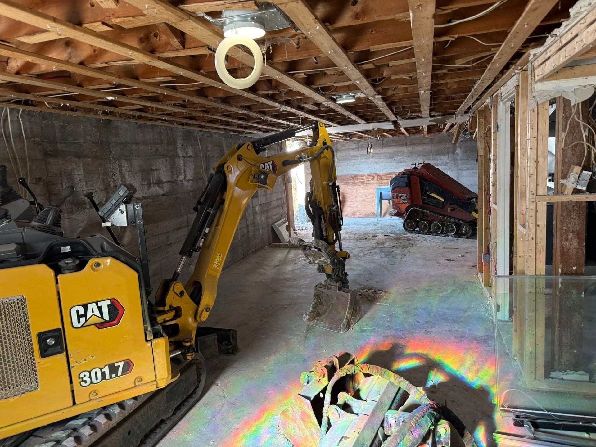 Mini-pelle jaune CAT sur un chantier de construction de sous-sol avec des débris et des poutres de plafond apparentes.