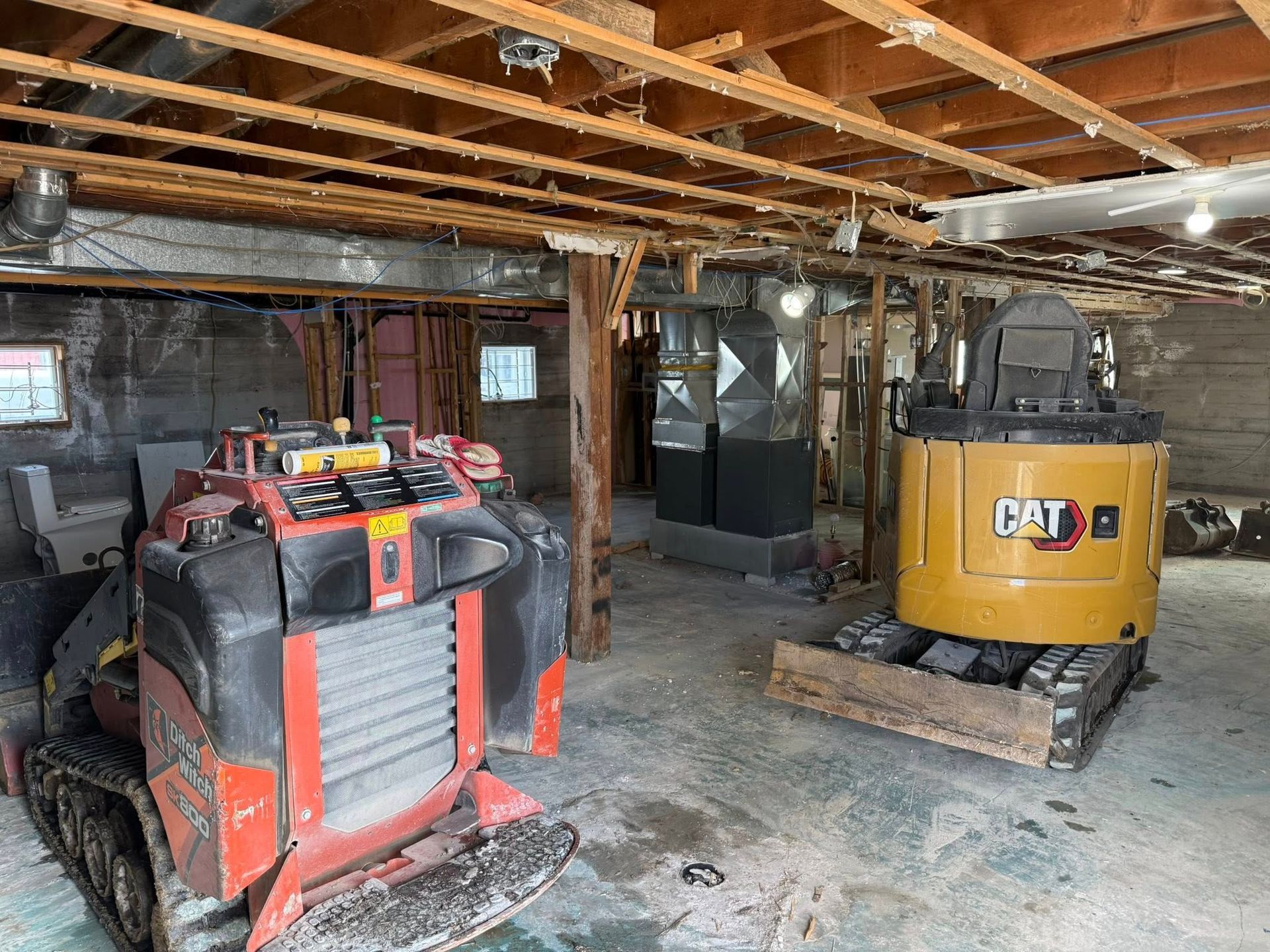 Sous-sol avec deux engins de chantier : un véhicule utilitaire rouge et une excavatrice CAT jaune sous des poutres apparentes.