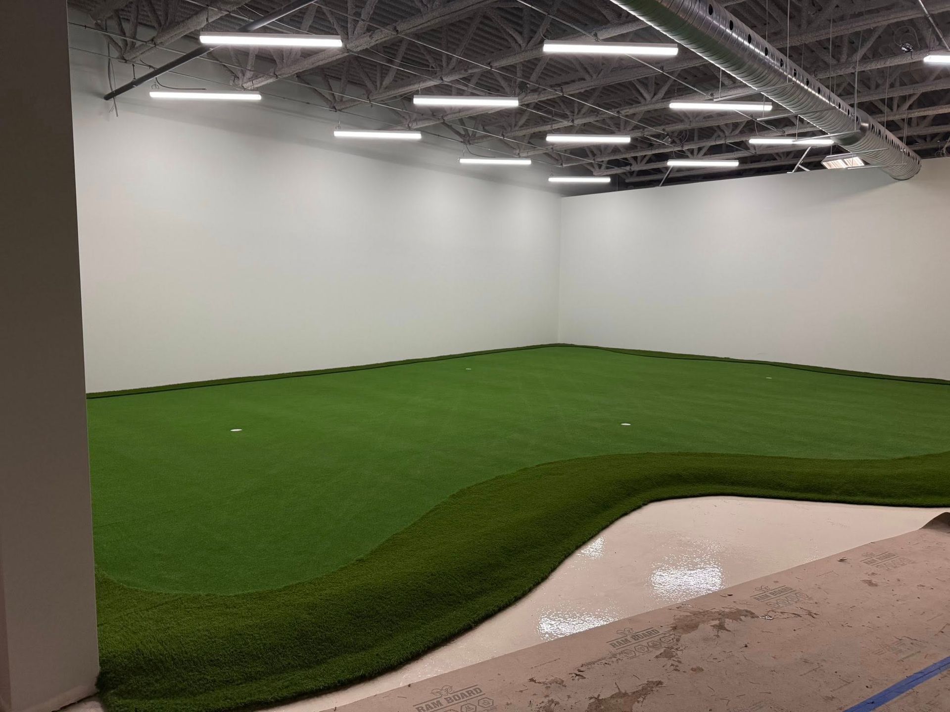 Un putting green intérieur dans une grande pièce blanche éclairée par des projecteurs et bordé d'un bunker.