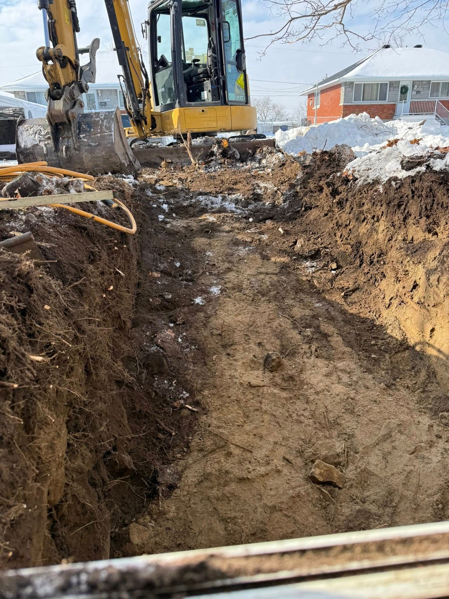 Une excavatrice creuse une profonde tranchée sur un chantier enneigé, avec des tas de terre de chaque côté.