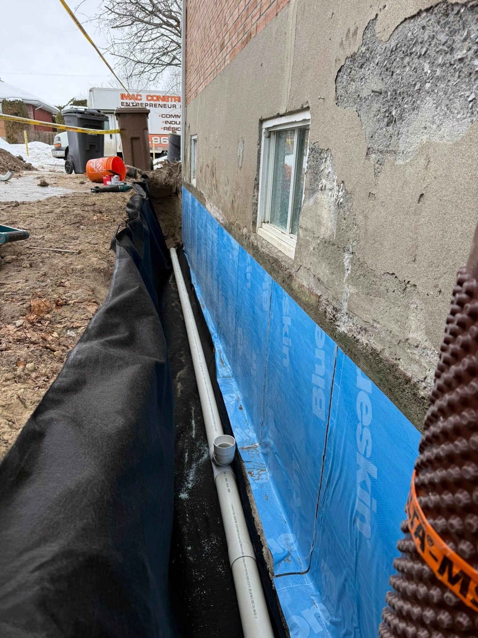 Chantier de construction avec bâche de fondation bleue, tranchée de gravier et mur de maison avec fenêtre