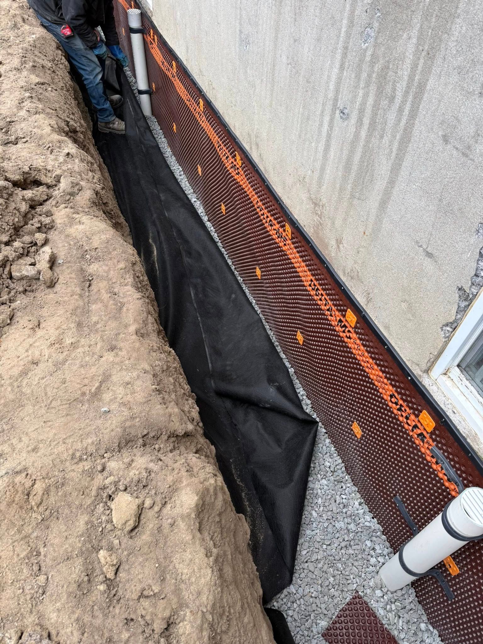 Tranchée le long d'un mur en béton avec barrière en tissu noir et en filet orange lors de travaux de drainage ou de construction