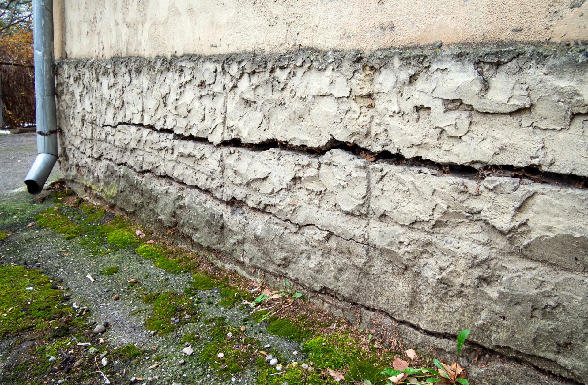 Mur en béton fissuré et altéré par les intempéries, jouxtant un chemin de gravier moussu et une descente de gouttière.
