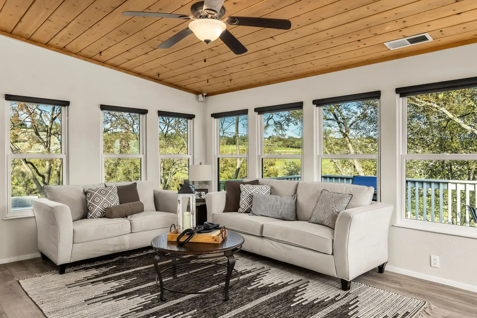 Sunlit sunroom with wood-paneled ceiling, large windows, ceiling fan, and neutral lounge seating.