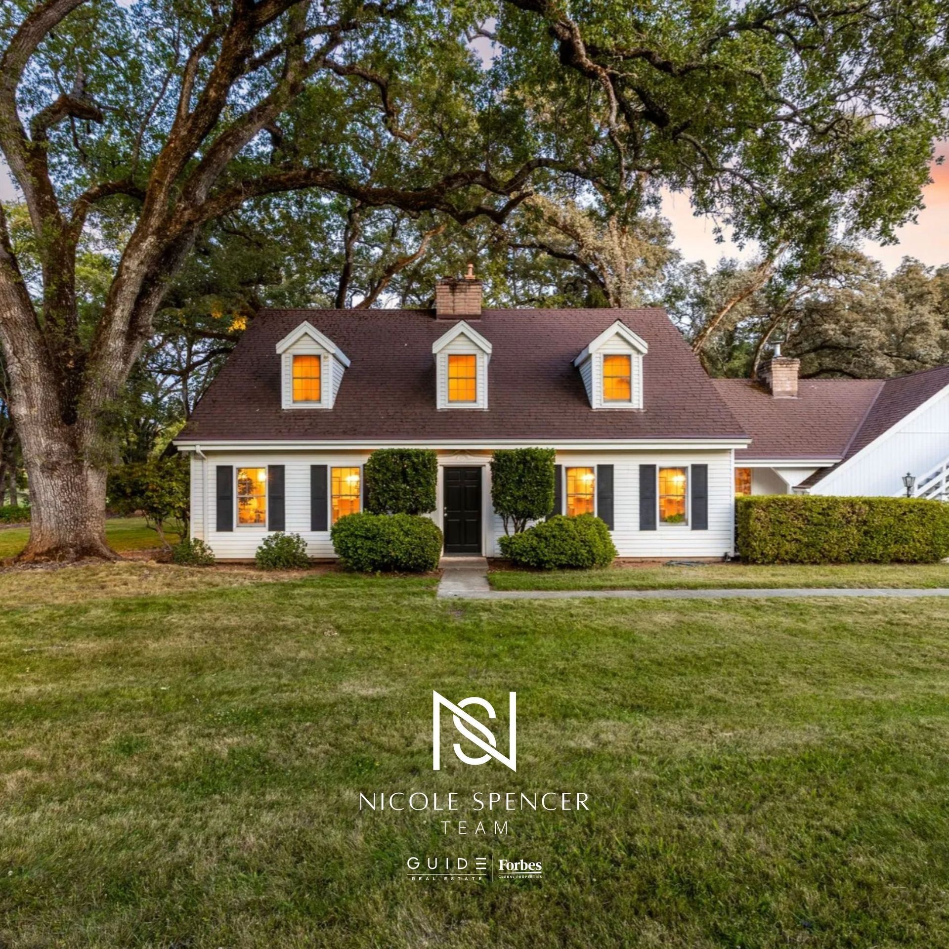 A charming white house with glowing windows nestled under large oak trees during a sunset, sold by Nicole Spencer.
