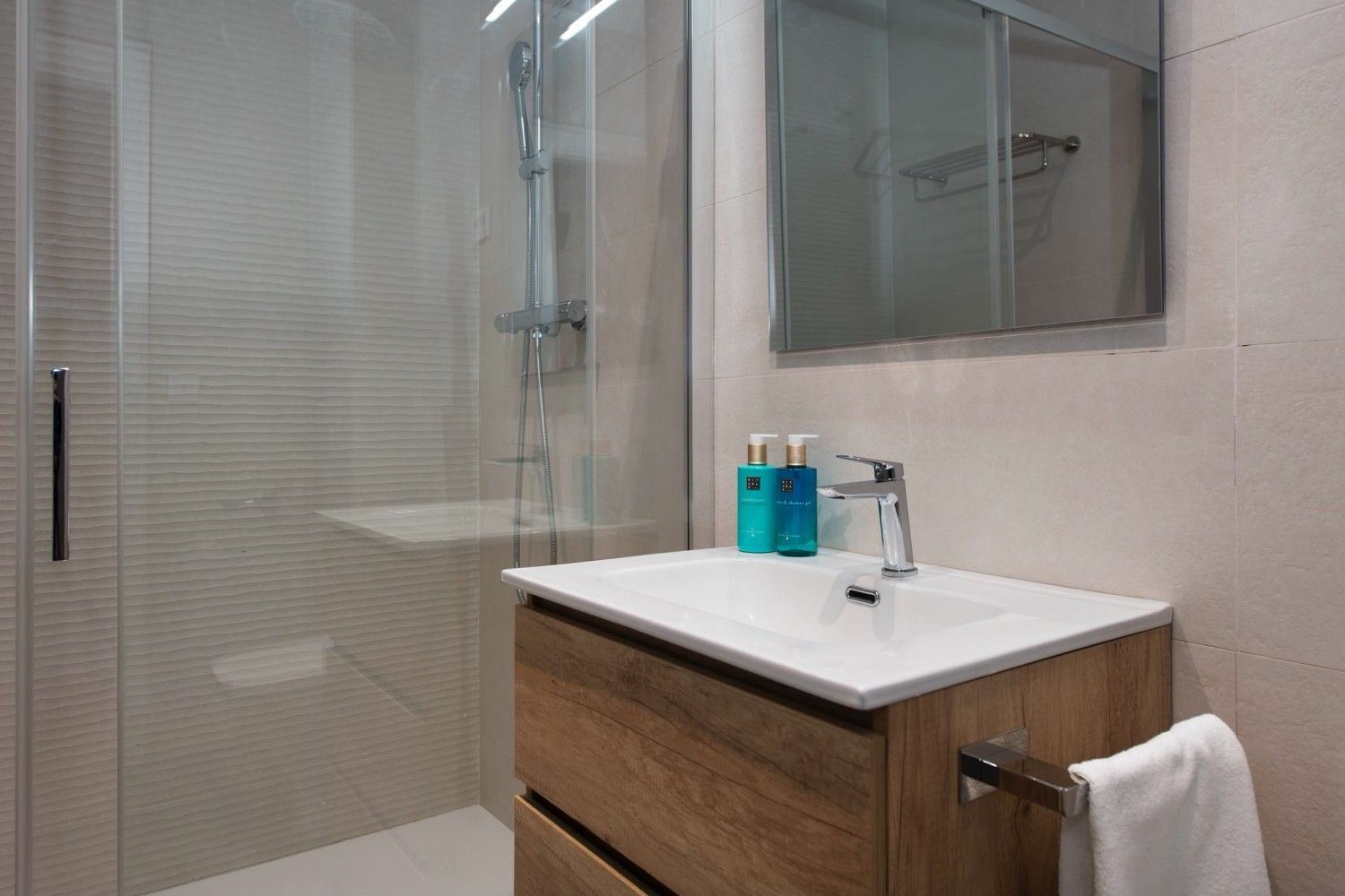 Baño moderno con ducha de cristal, lavabo con tocador, espejo y dispensadores de jabón azules.